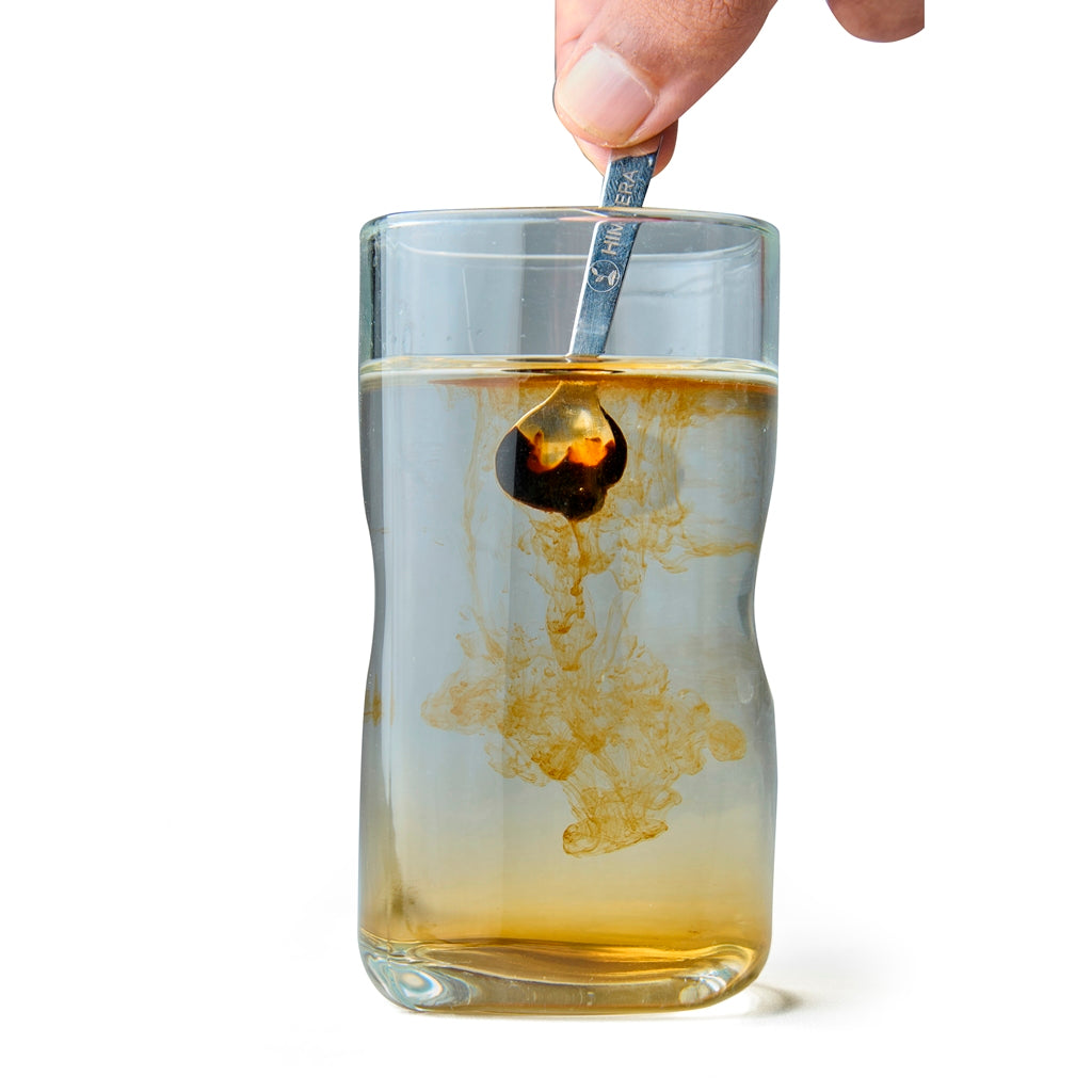 Glass of amber liquid with a spoon stirring it, on a white background